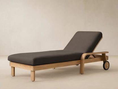 Zoe Outdoor Chaise with teak wood frame, dark gray cushion, and wheels, on a stone patio against a beige wall.