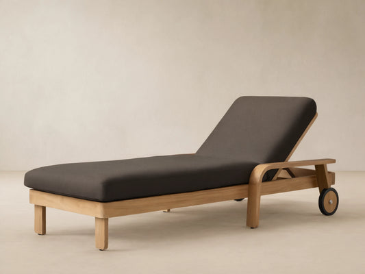 Zoe Outdoor Chaise with teak wood frame, dark gray cushion, and wheels, on a stone patio against a beige wall.