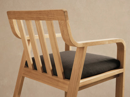 Zoe Outdoor Dining Arm Chair, light teak wood with slatted back, straight arms, and a dark gray seat cushion, front view.