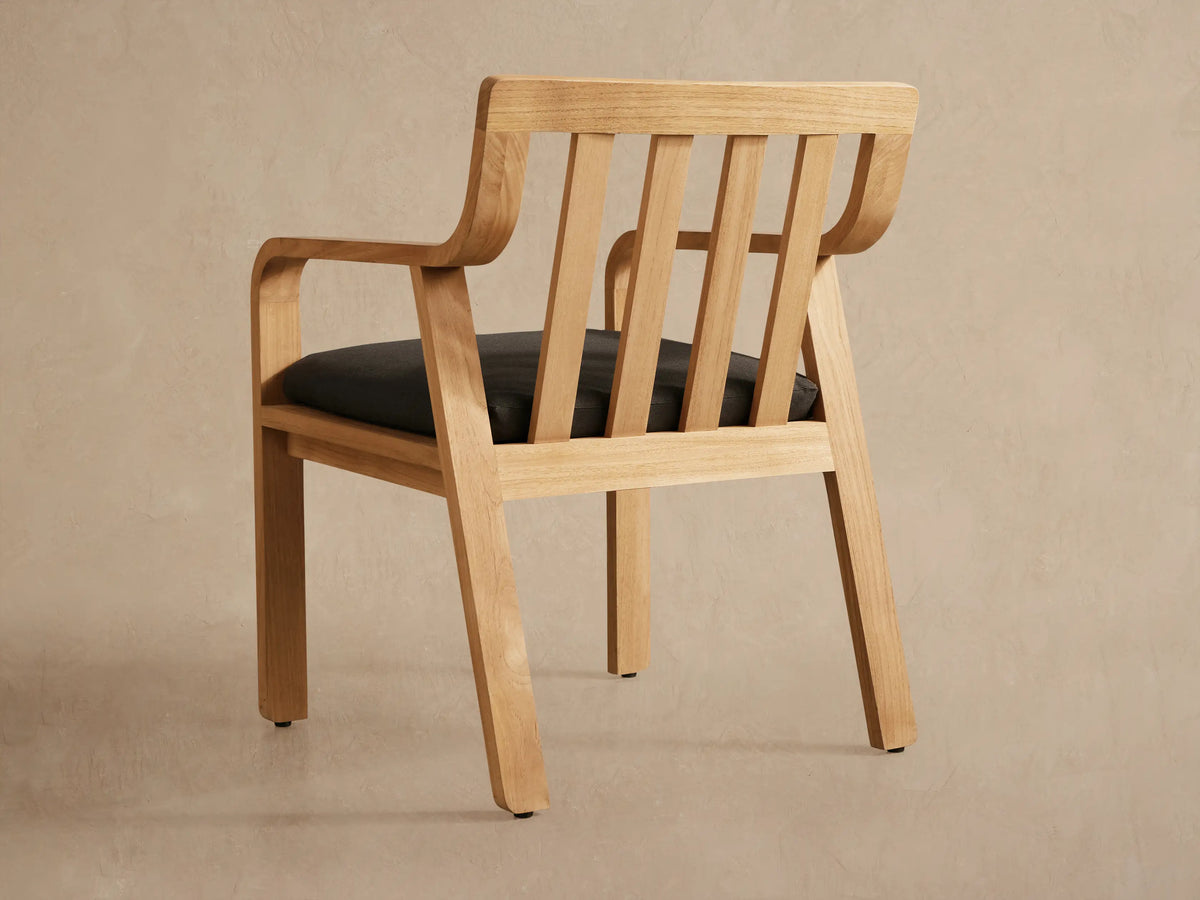 Close-up of a Zoe Outdoor Dining Arm Chair, showing its natural teak wood frame, vertical slat back, curved armrests, and dark gray seat cushion, angled slightly right.