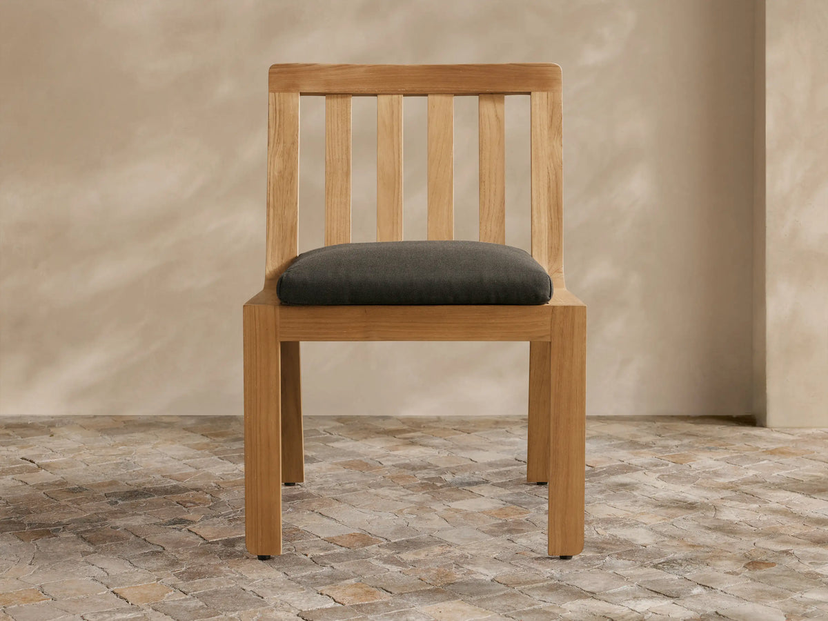 Light teak Zoe Outdoor Dining Side Chair with slatted back and dark gray cushion, angled slightly right on a stone floor.