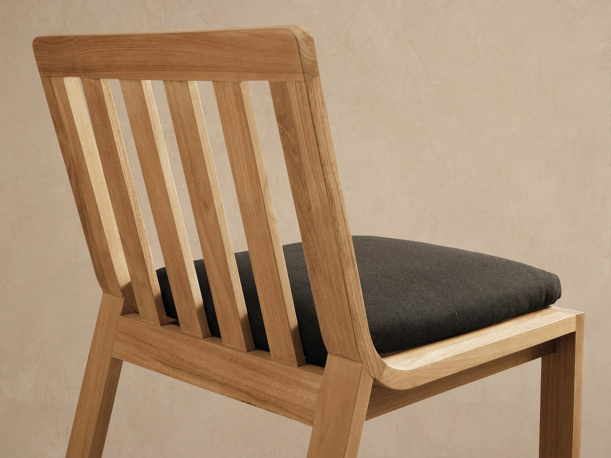 Zoe Outdoor Dining Side Chair, light teak wood with slatted back, dark gray cushion, facing forward on stone tiled floor.
