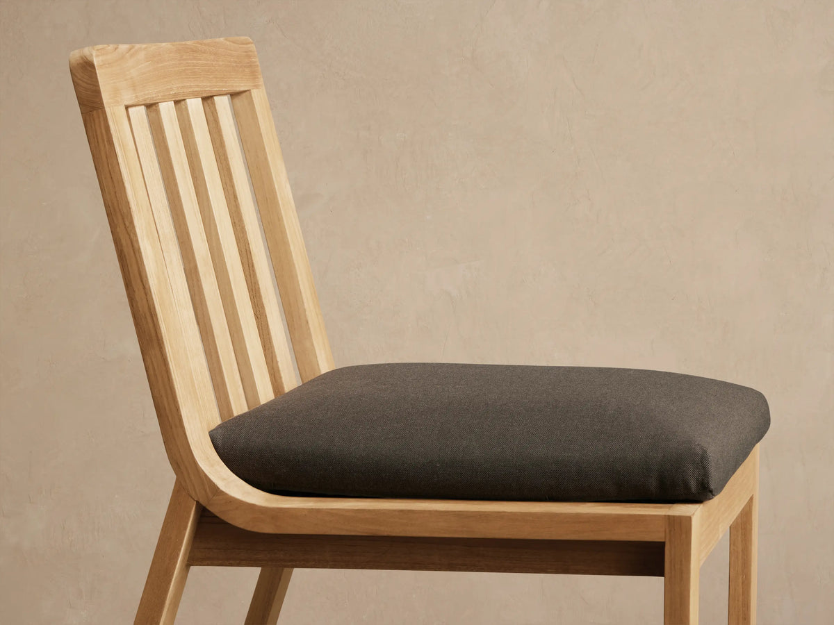 Zoe Outdoor Dining Side Chair, natural teak wood frame with vertical slatted back and dark grey cushion, angled right.
