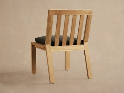 Close-up of Zoe Outdoor Dining Side Chair, natural teak wood, slatted back, dark gray fabric cushion, angled view.