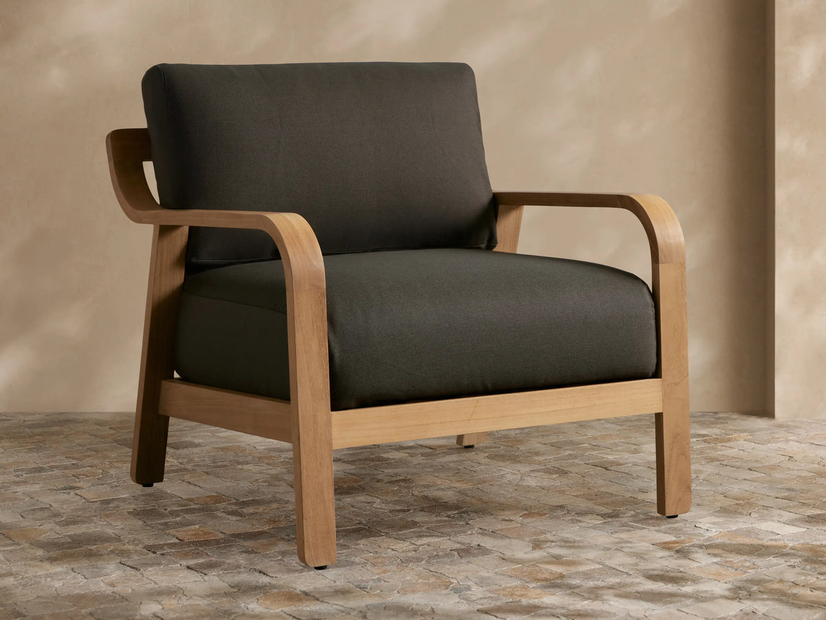 Zoe Outdoor Lounge Chair: Light teak wood frame with dark grey cushions, curved armrests, on a rustic stone floor.