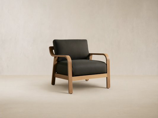 Zoe Outdoor Lounge Chair: Light teak wood frame with dark grey cushions, curved armrests, on a rustic stone floor.