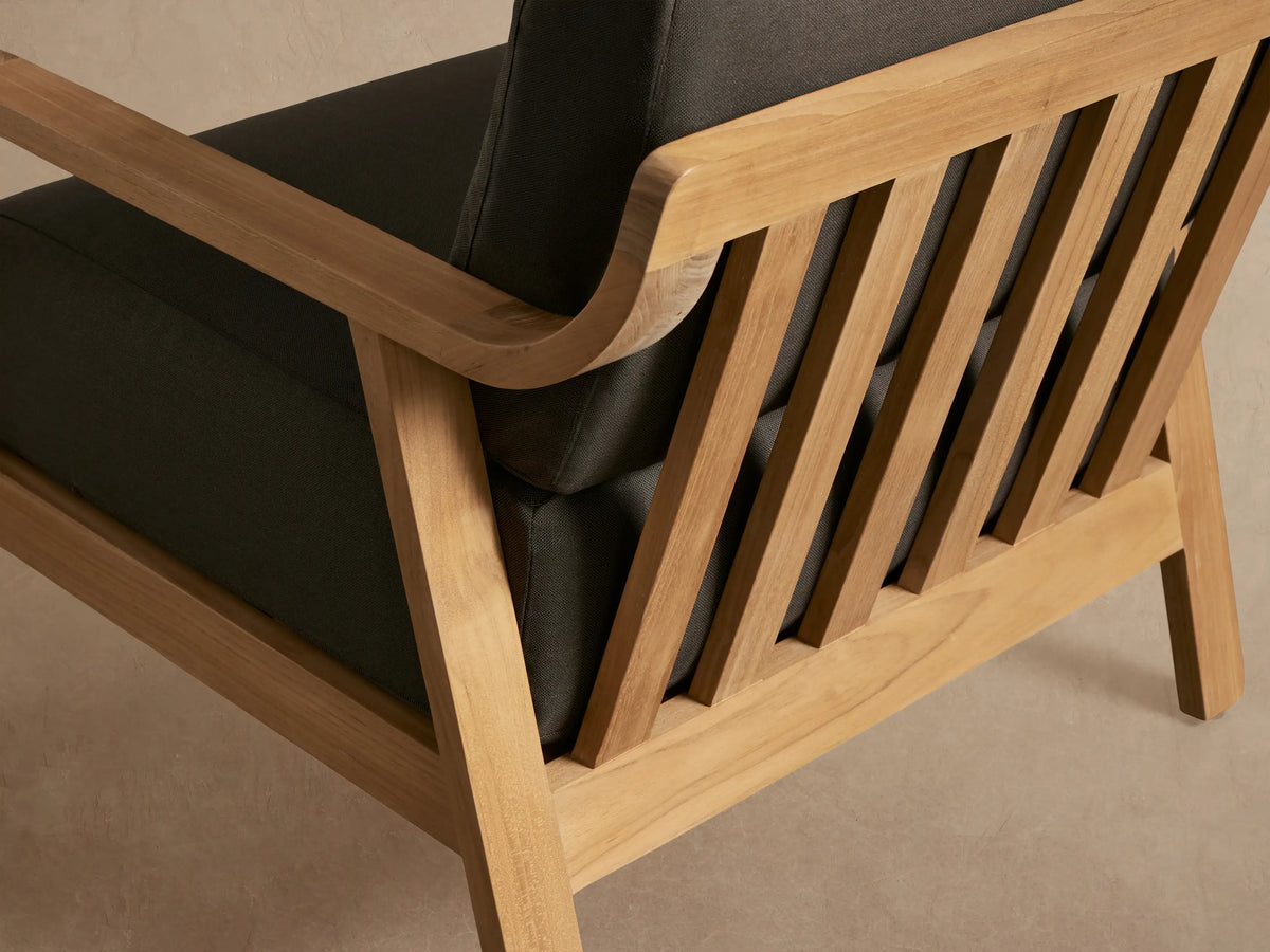 Zoe Outdoor Lounge Chair with a warm, natural teak wood frame and thick dark gray cushions, viewed from the front.