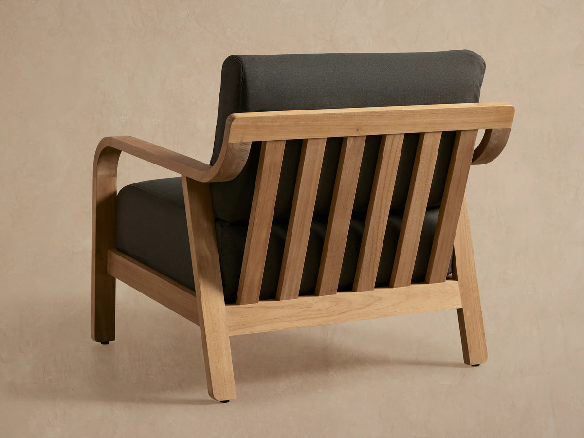 Angled close-up of Zoe Outdoor Lounge Chair. Golden teak wood frame with slatted back and dark gray seat and back cushions.