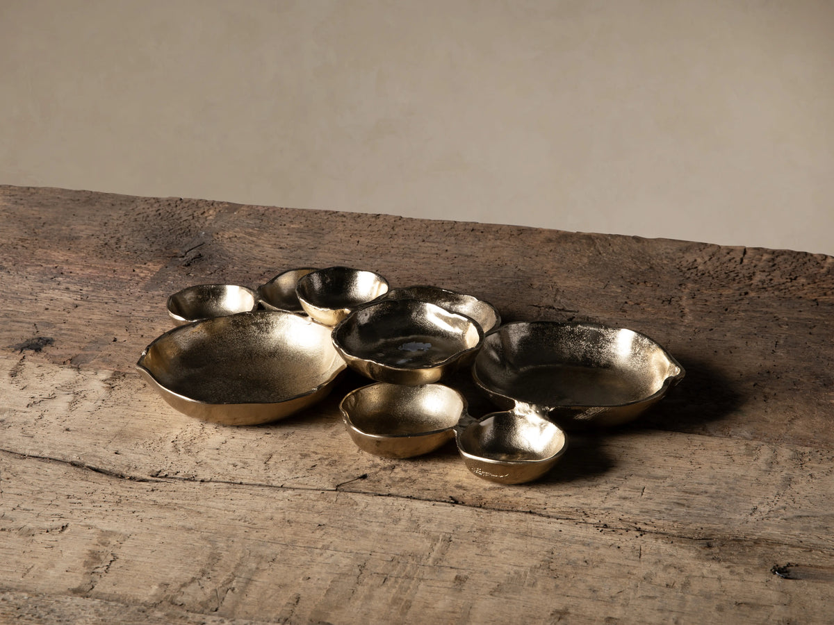A group of Klasa dark antiqued gold aluminum platters and bowls of various sizes, with wavy edges, on a rustic wooden table.