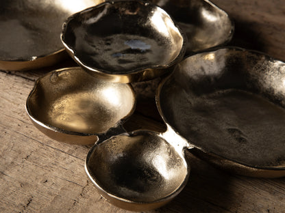 A group of Klasa dark antiqued gold aluminum platters and bowls of various sizes, with wavy edges, on a rustic wooden table.