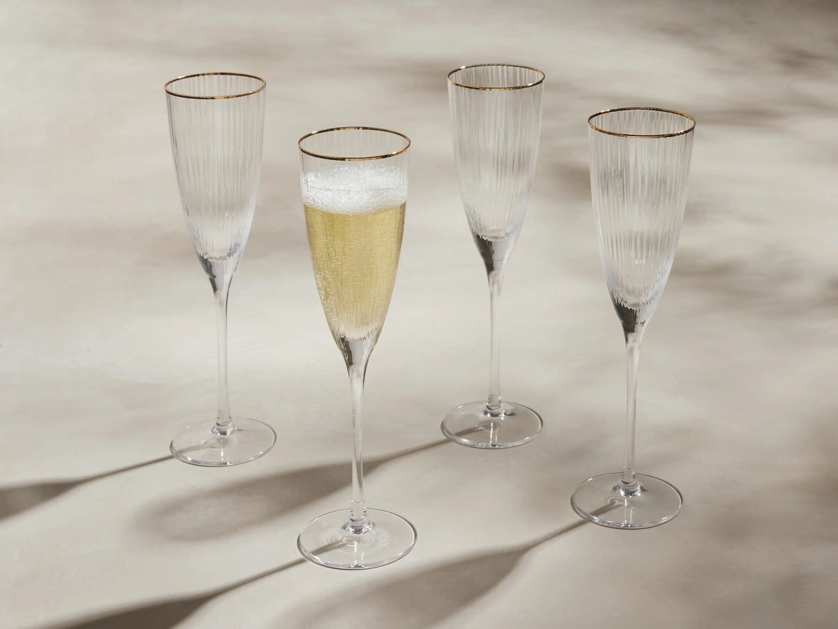 Four Anisa ribbed champagne flutes with gold rims stand. One filled with bubbly champagne, on a light surface with long shadows.