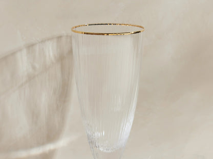 Four Anisa ribbed champagne flutes with gold rims stand. One filled with bubbly champagne, on a light surface with long shadows.