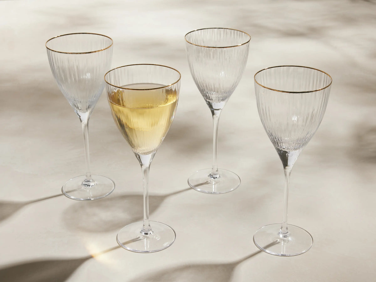 Set of 4 Anisa ribbed, hand-blown wine glasses with gold rims, one filled with yellow liquid, on a light surface.
