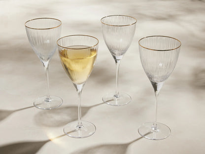 Set of 4 Anisa ribbed, hand-blown wine glasses with gold rims, one filled with yellow liquid, on a light surface.