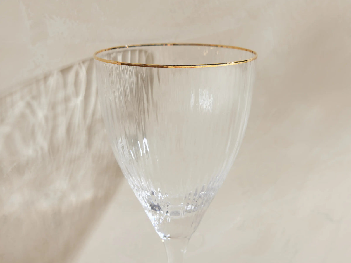 Set of 4 Anisa ribbed, hand-blown wine glasses with gold rims, one filled with yellow liquid, on a light surface.