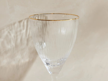 Set of 4 Anisa ribbed, hand-blown wine glasses with gold rims, one filled with yellow liquid, on a light surface.