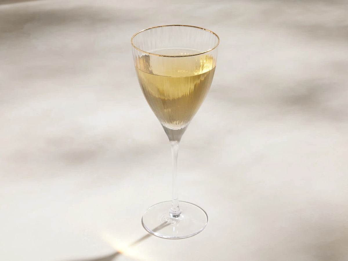 Close-up of a clear Anisa Glassware goblet with ribbed texture and a gold rim, casting light reflections on a beige wall.