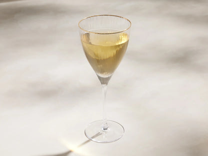 Close-up of a clear Anisa Glassware goblet with ribbed texture and a gold rim, casting light reflections on a beige wall.