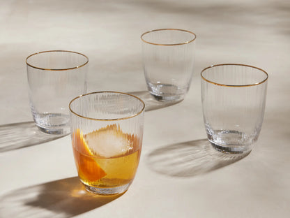 Four clear, ribbed Anisa Double Old Fashioned Glasses with gold rims. One contains an amber drink with ice and orange peel.