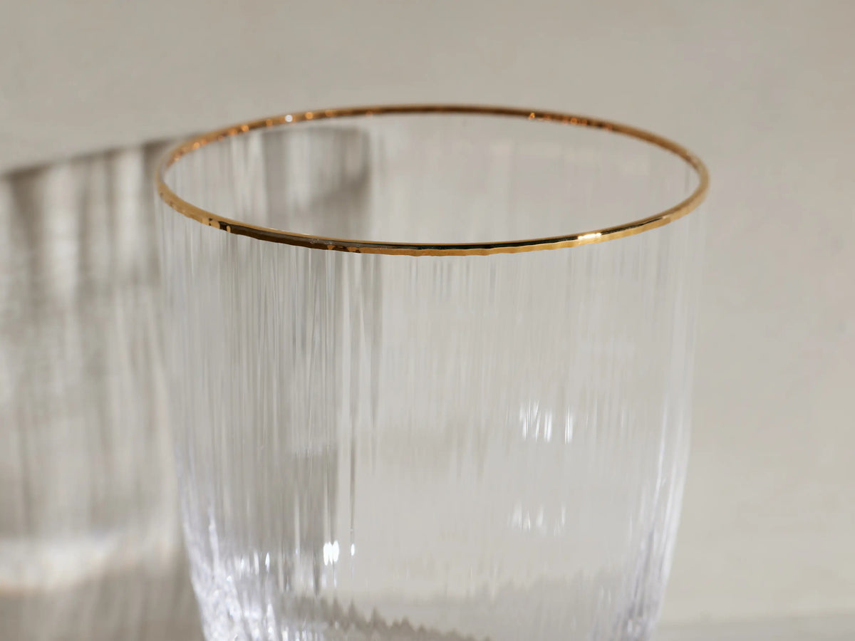 Four clear, ribbed Anisa Double Old Fashioned Glasses with gold rims. One contains an amber drink with ice and orange peel.