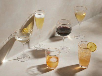Hand-blown, ribbed Anisa Double Old Fashioned glass with hand-painted gold rim, amber drink, ice, and orange peel.