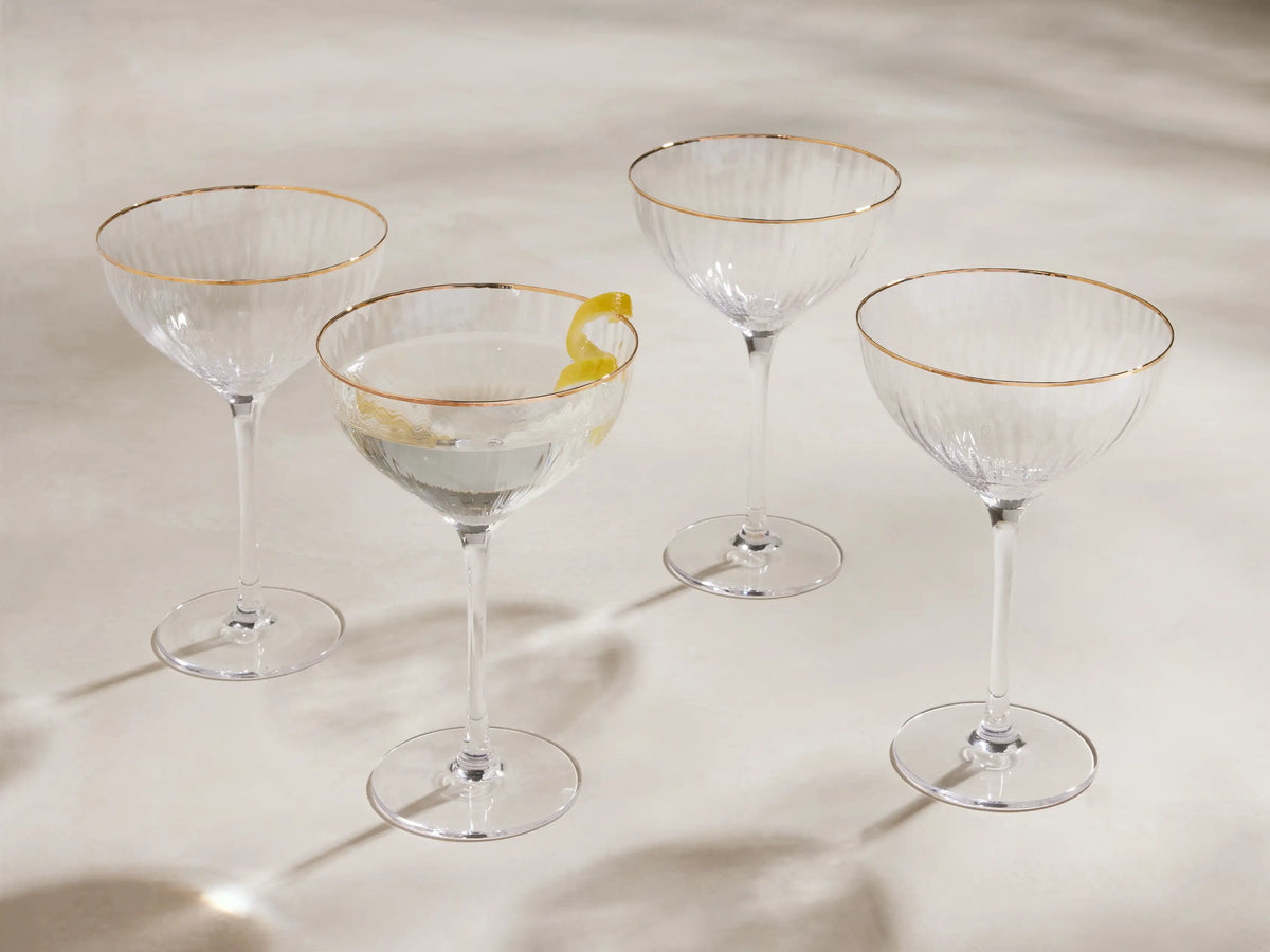 Four Anisa ribbed cocktail glasses with gold rims, one filled with liquid & lemon peel, on a light surface.