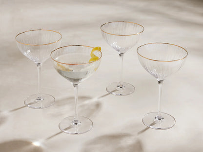 Four Anisa ribbed cocktail glasses with gold rims, one filled with liquid & lemon peel, on a light surface.