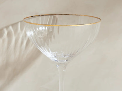 Four Anisa ribbed cocktail glasses with gold rims, one filled with liquid & lemon peel, on a light surface.