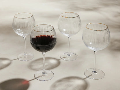 Front view of four clear Anisa Red Wine Glasses with ribbed bowls and gold rims, one filled with red wine.