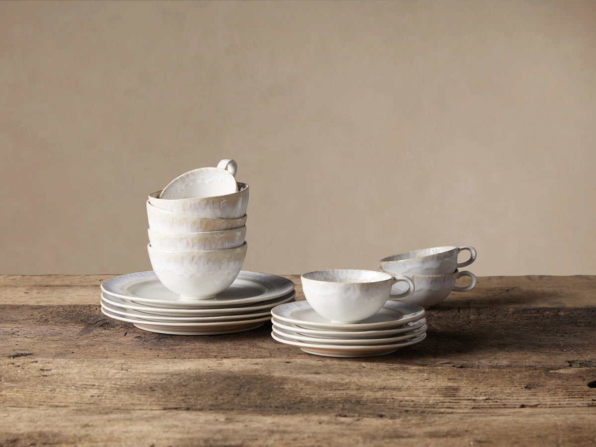 Bela Dinnerware set: Stacked creamy off-white, glazed stoneware bowls & plates, with mugs & saucers on a wooden table.