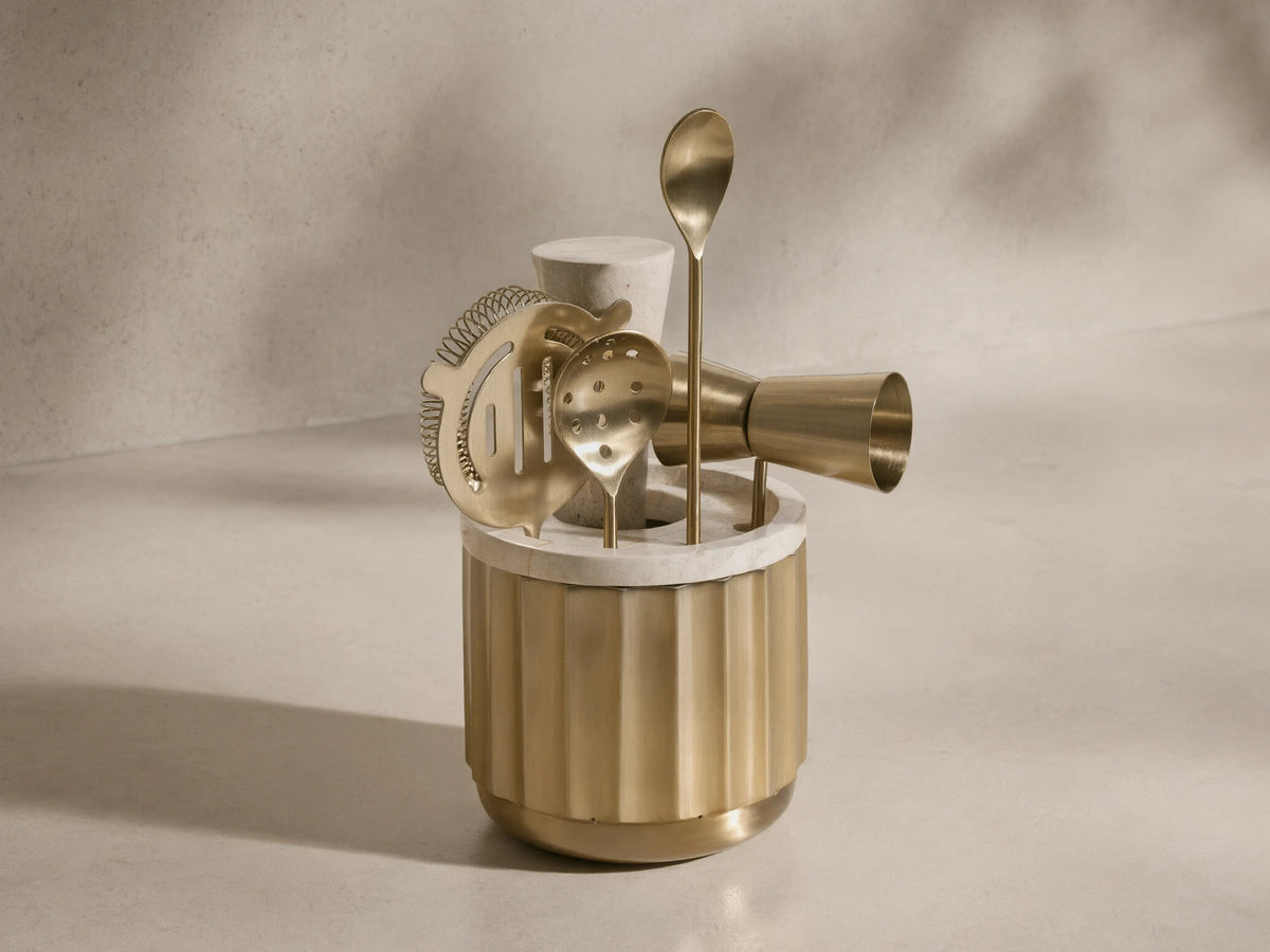 Front view of the golden Dayan Bar Tools set, including a fluted brass holder with a white marble top, strainer, jigger, and spoons.