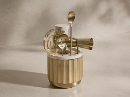 Front view of the golden Dayan Bar Tools set, including a fluted brass holder with a white marble top, strainer, jigger, and spoons.