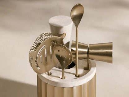 Front view of the golden Dayan Bar Tools set, including a fluted brass holder with a white marble top, strainer, jigger, and spoons.
