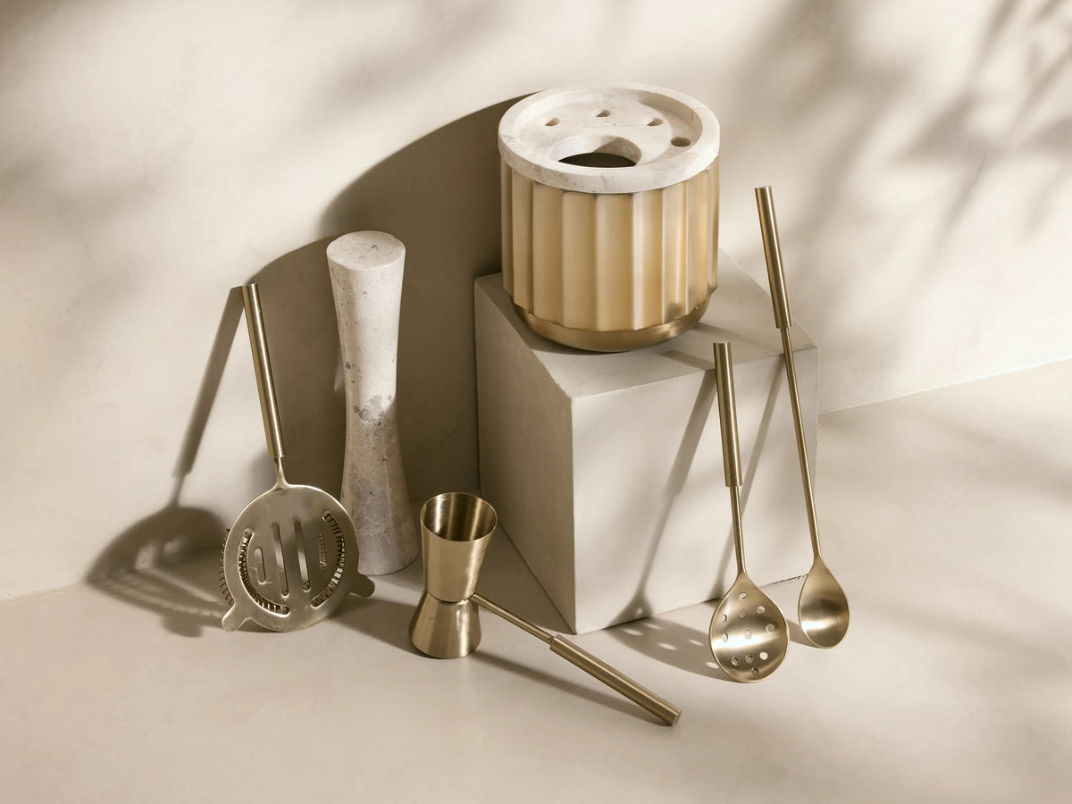 Close-up of brushed gold Dayan Bar Tools: strainer, spoon, jigger, and marble muddler in a ribbed gold holder.