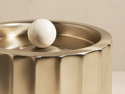 Fluted brass Dayana Ice Bucket holding a green champagne bottle with ice. Its lid, with a white marble handle, rests beside it.