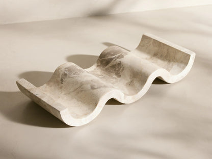 Angled view of the wavy, creamy white Dayana Wine Rack, crafted from natural marble with gray veining, holding a wine bottle.