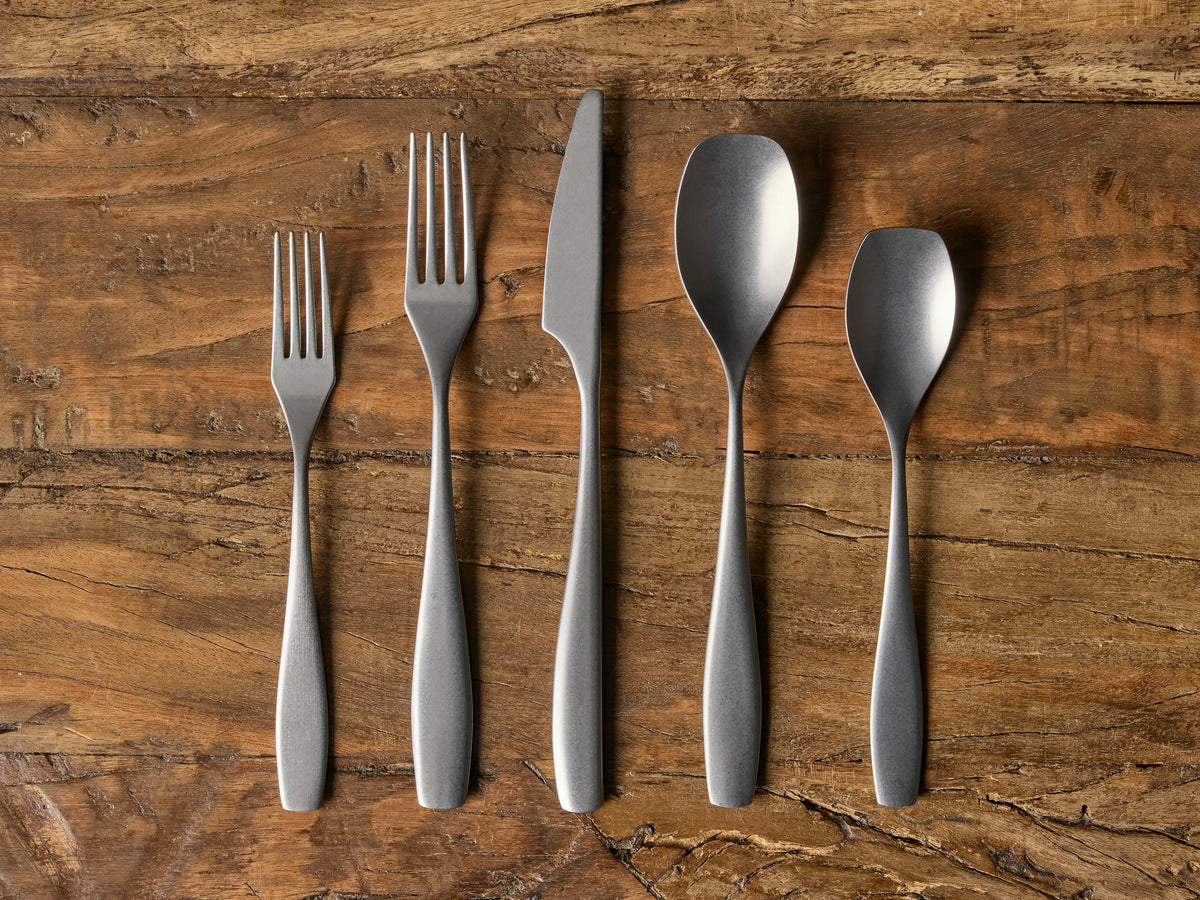 Esma Five Piece Place Setting includes knife, two forks, and two spoons in stonewash finish, arranged on a wooden surface.