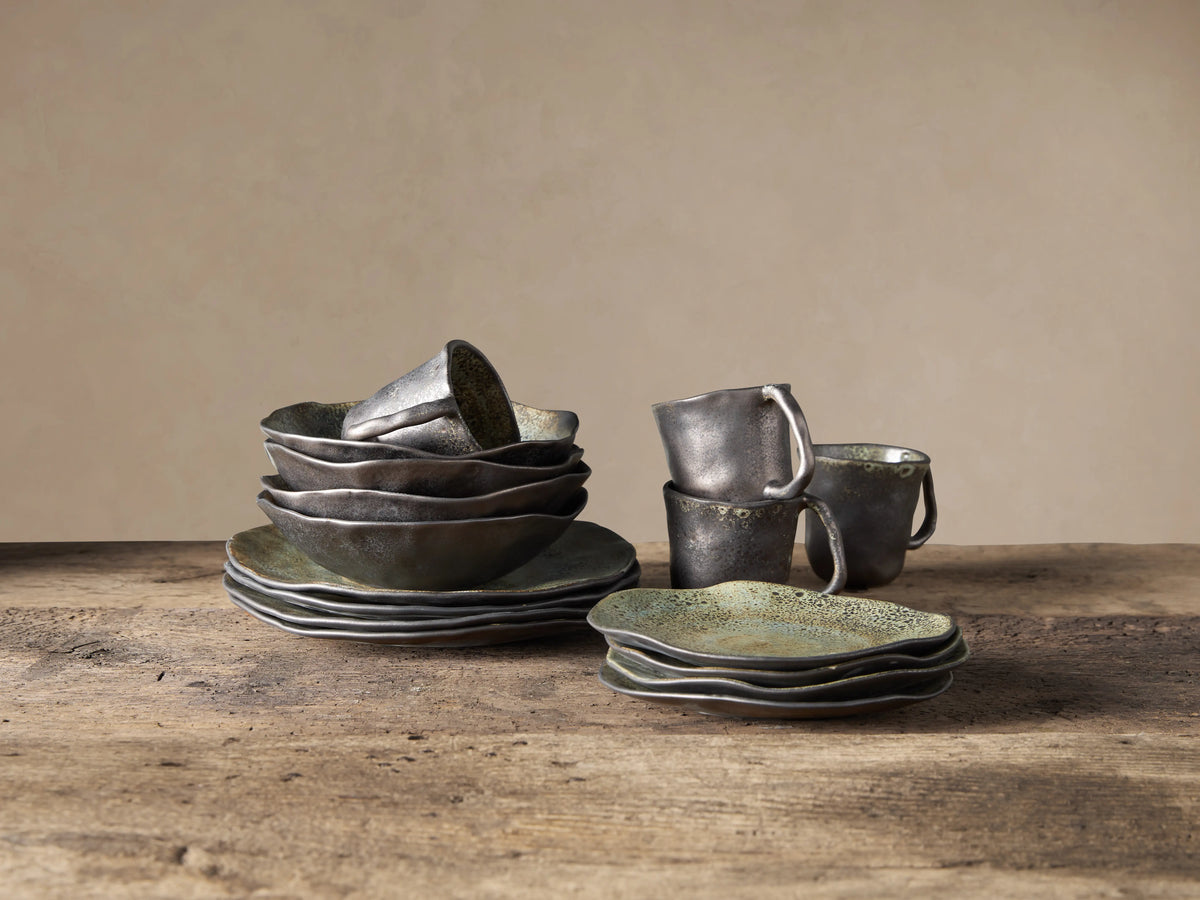 Gallatin Dinnerware 16-piece set: stacked irregular-edged bowls, plates, mugs with mottled green-brown reactive glaze on wood.
