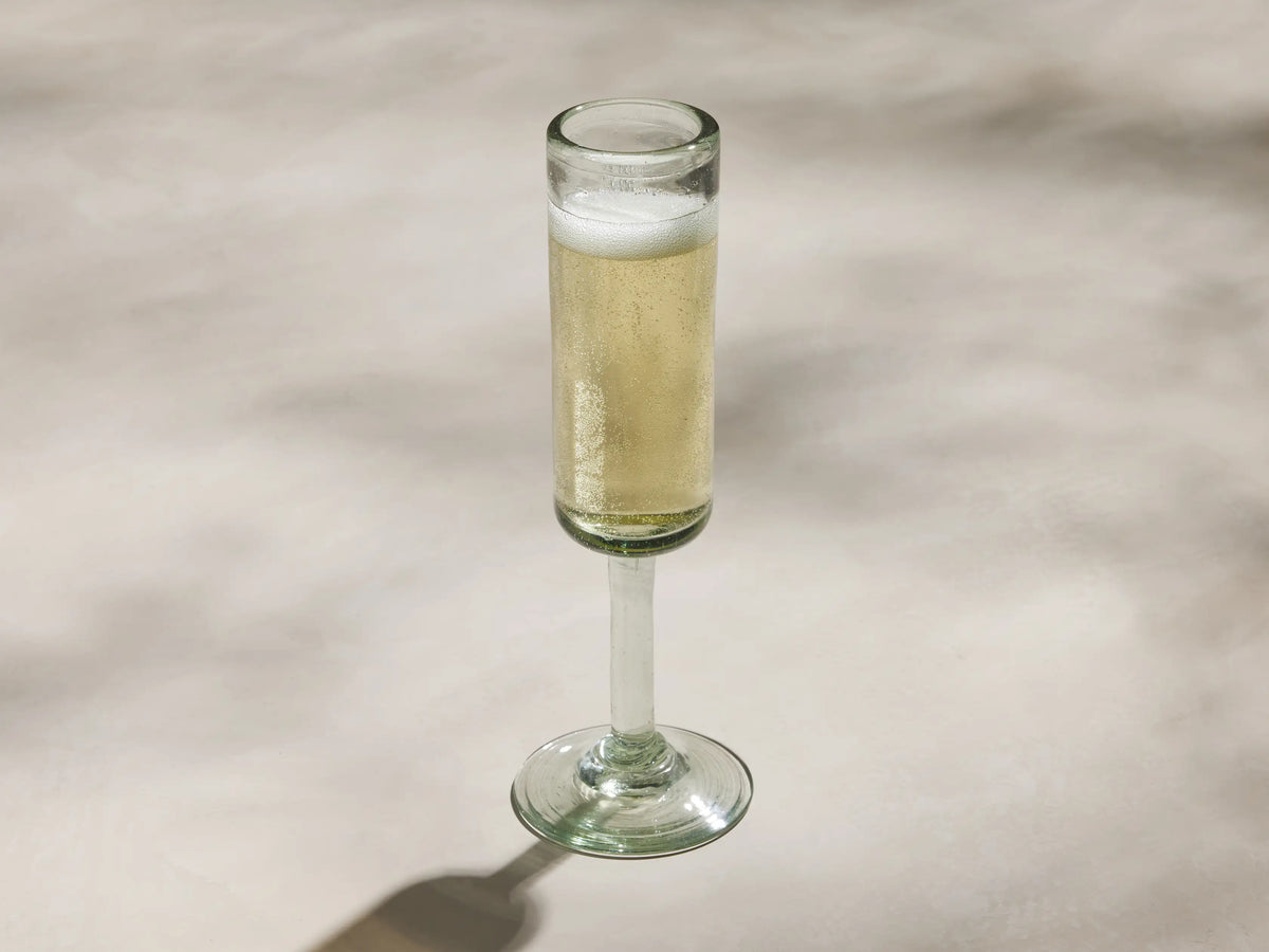 Single, upright, clear Granada Champagne Glass. Hand-blown recycled glass with a subtle green tint and imperfections, against a light wall.