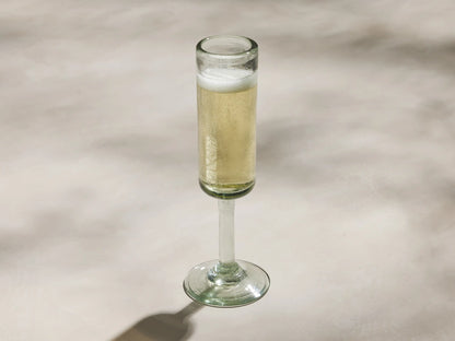 Single, upright, clear Granada Champagne Glass. Hand-blown recycled glass with a subtle green tint and imperfections, against a light wall.