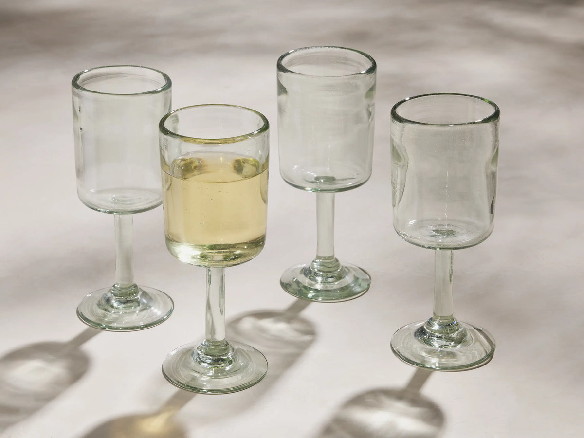 Four Granada recycled glass wine glasses, cylindrical and stemmed. One holds yellow wine, all cast shadows on a light surface.