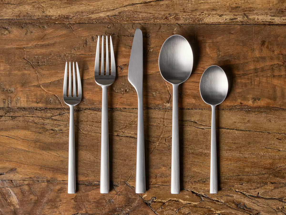 Holmes Five Piece Place Setting: Matte stainless steel forks, knife, and spoons arranged vertically on a rustic wooden table.