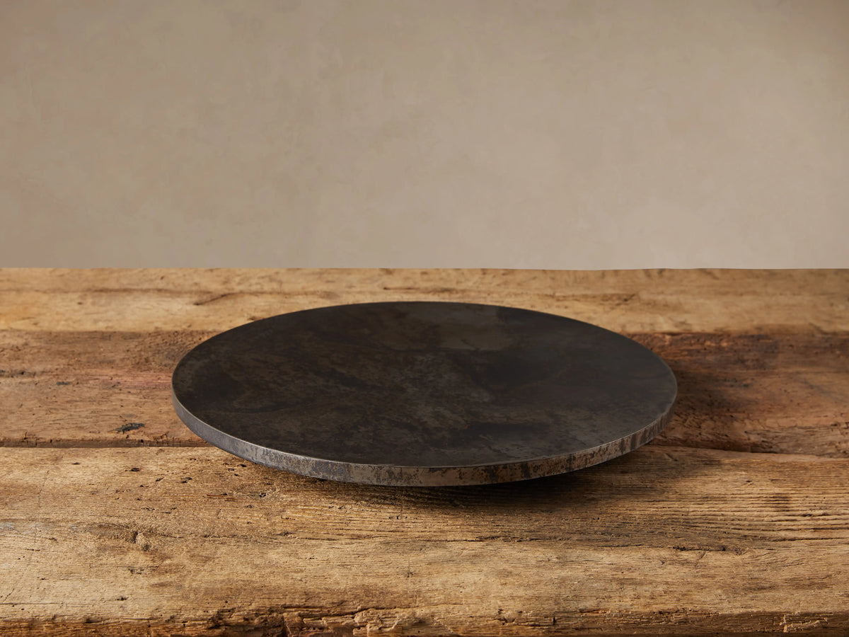 Dark, round Sur Metal Lazy Susan with an oxidized finish, resting on a rustic wooden table. Beige wall in background.