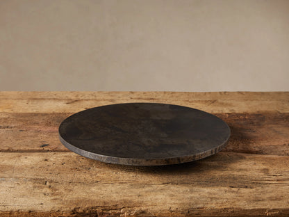 Dark, round Sur Metal Lazy Susan with an oxidized finish, resting on a rustic wooden table. Beige wall in background.