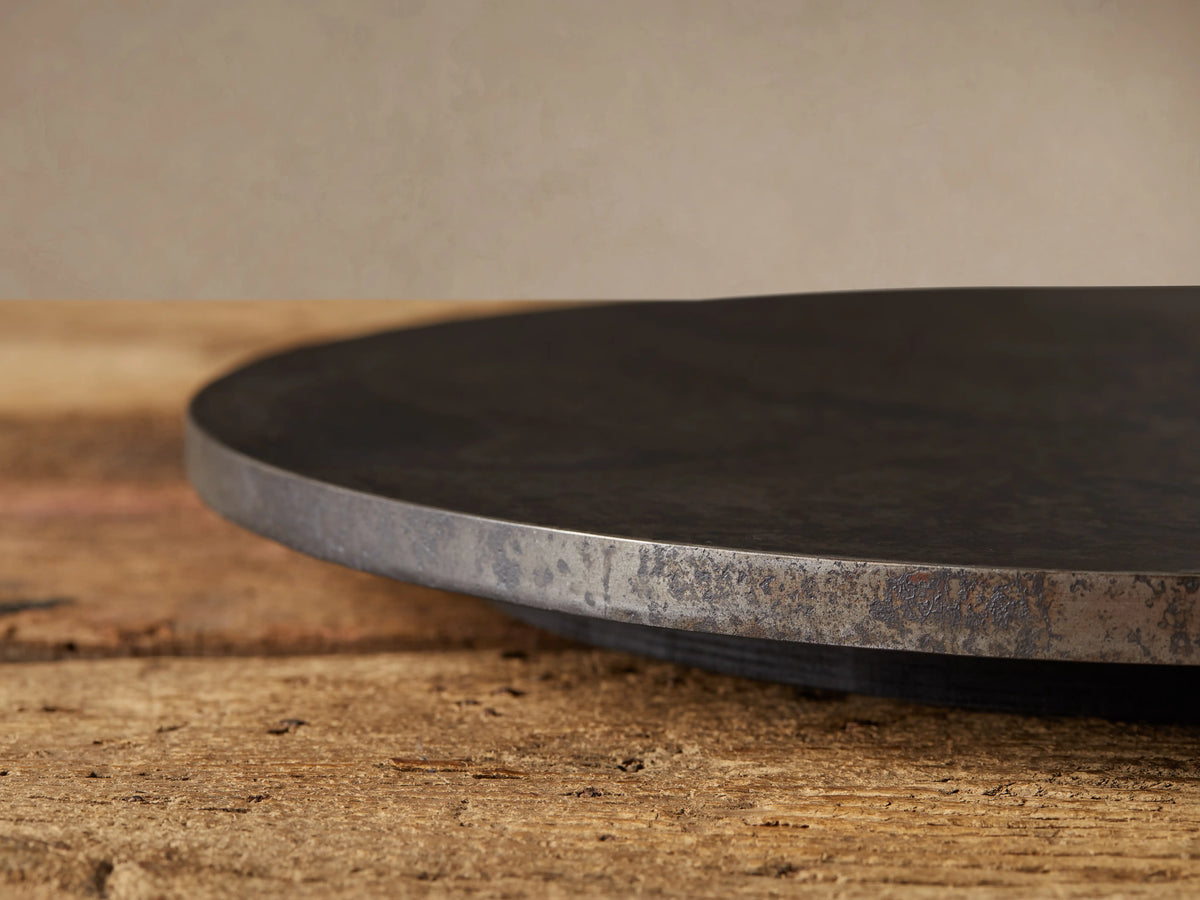 Dark, round Sur Metal Lazy Susan with an oxidized finish, resting on a rustic wooden table. Beige wall in background.
