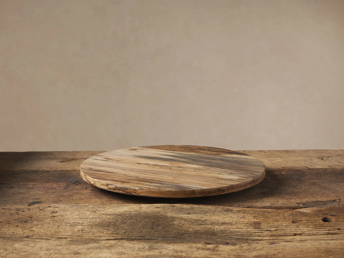 Round Madeira Natural Lazy Susan, crafted from varied light & dark reclaimed wood, rests on a rustic wood table against a beige wall.
