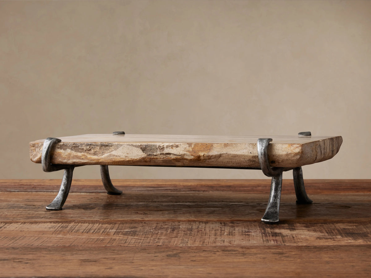 Petrified Wood Serving Board, light-toned with organic edges, held by dark gray wrought iron legs, sitting on a rustic wooden table.