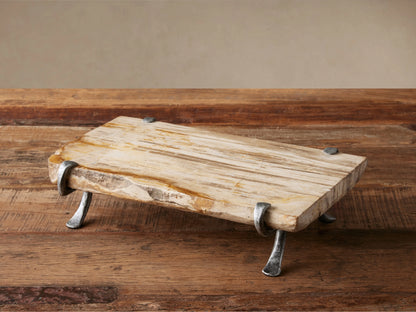 Petrified Wood Serving Board, light-toned with organic edges, held by dark gray wrought iron legs, sitting on a rustic wooden table.
