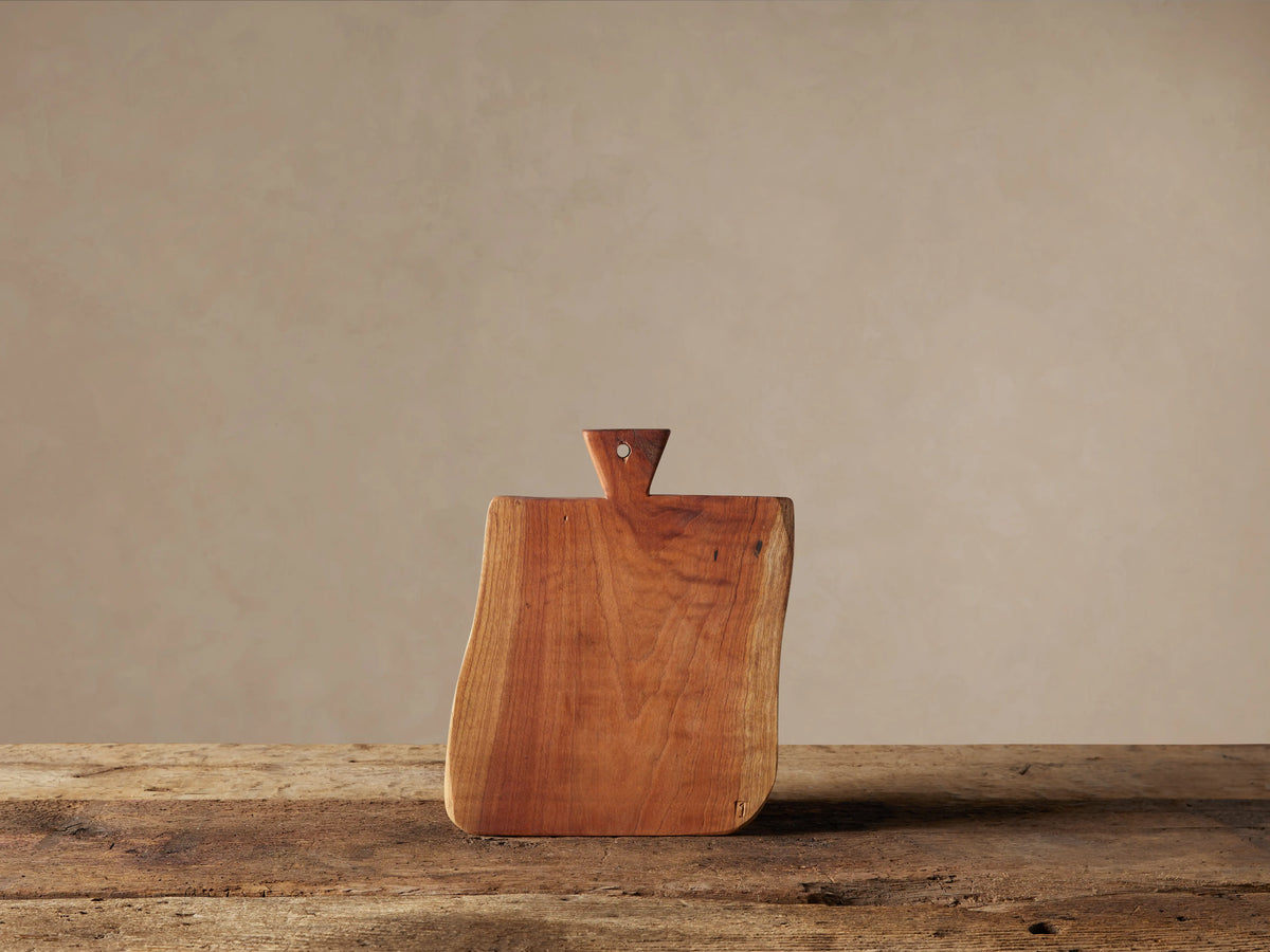 Reclaimed wood cutting board, warm reddish-brown with natural grain, standing upright on a rustic wooden table. It has an irregular shape and a small triangular handle with a hole.