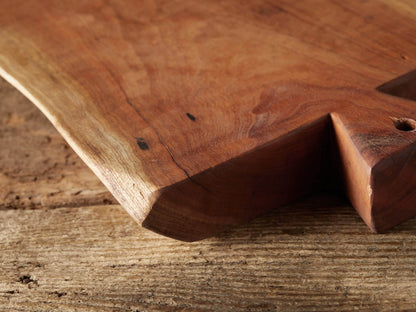 Reclaimed wood cutting board, warm reddish-brown with natural grain, standing upright on a rustic wooden table. It has an irregular shape and a small triangular handle with a hole.